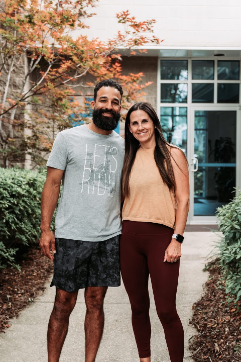 A smiling man and woman stand arm in arm outside a building with glass doors. They're surrounded by greenery and a tree with orange leaves, creating a warm, welcoming atmosphere.