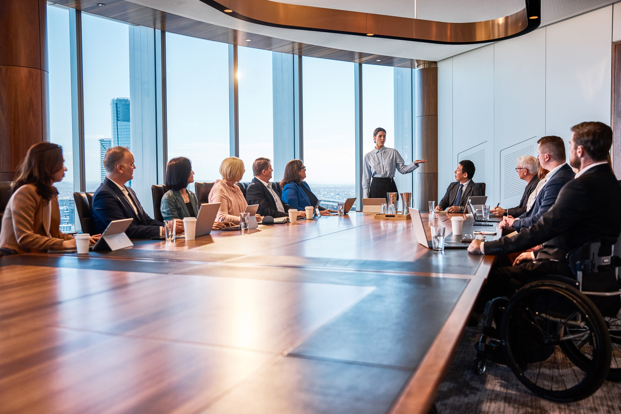 Business meeting in a modern boardroom with a long wooden table, where a person is standing and presenting to colleagues seated around, with large windows showing the Brisbane skyline.