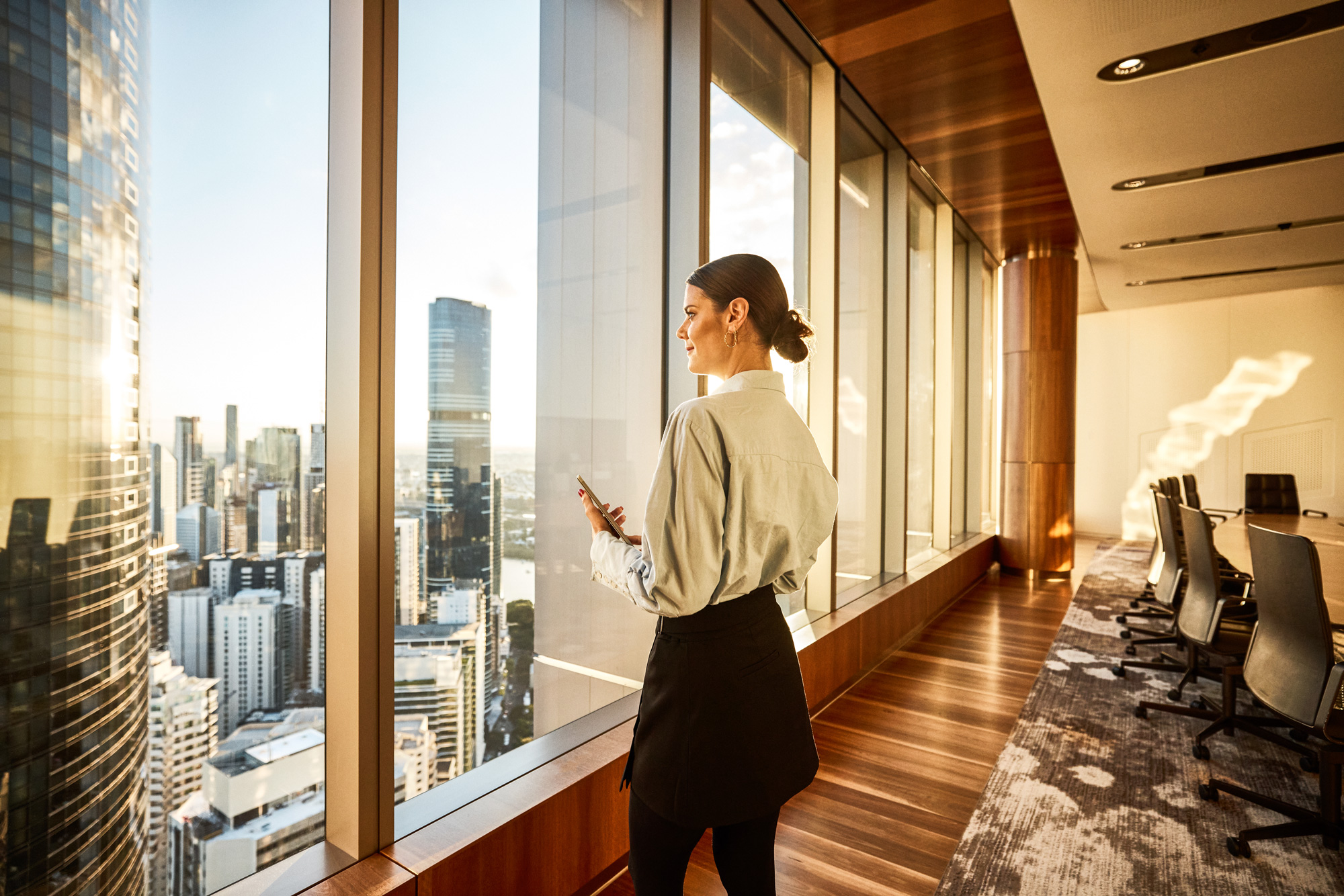 Woman standing in a modern high-rise office, looking out large windows over a city skyline while holding a smartphone.