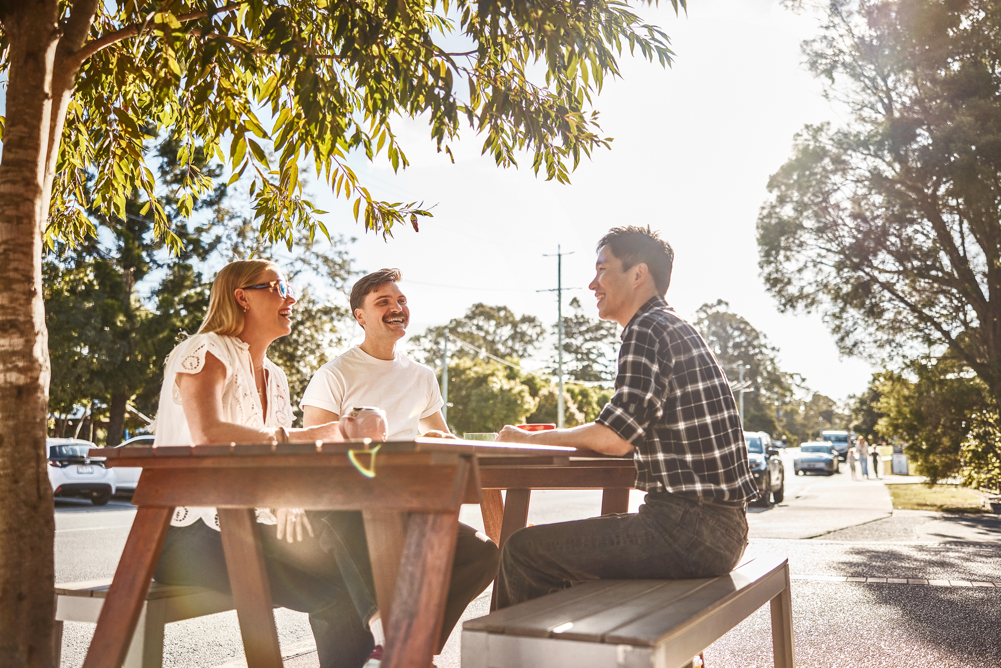 Three people sitting at a wooden picnic table under a tree, laughing and talking together on a sunny day.