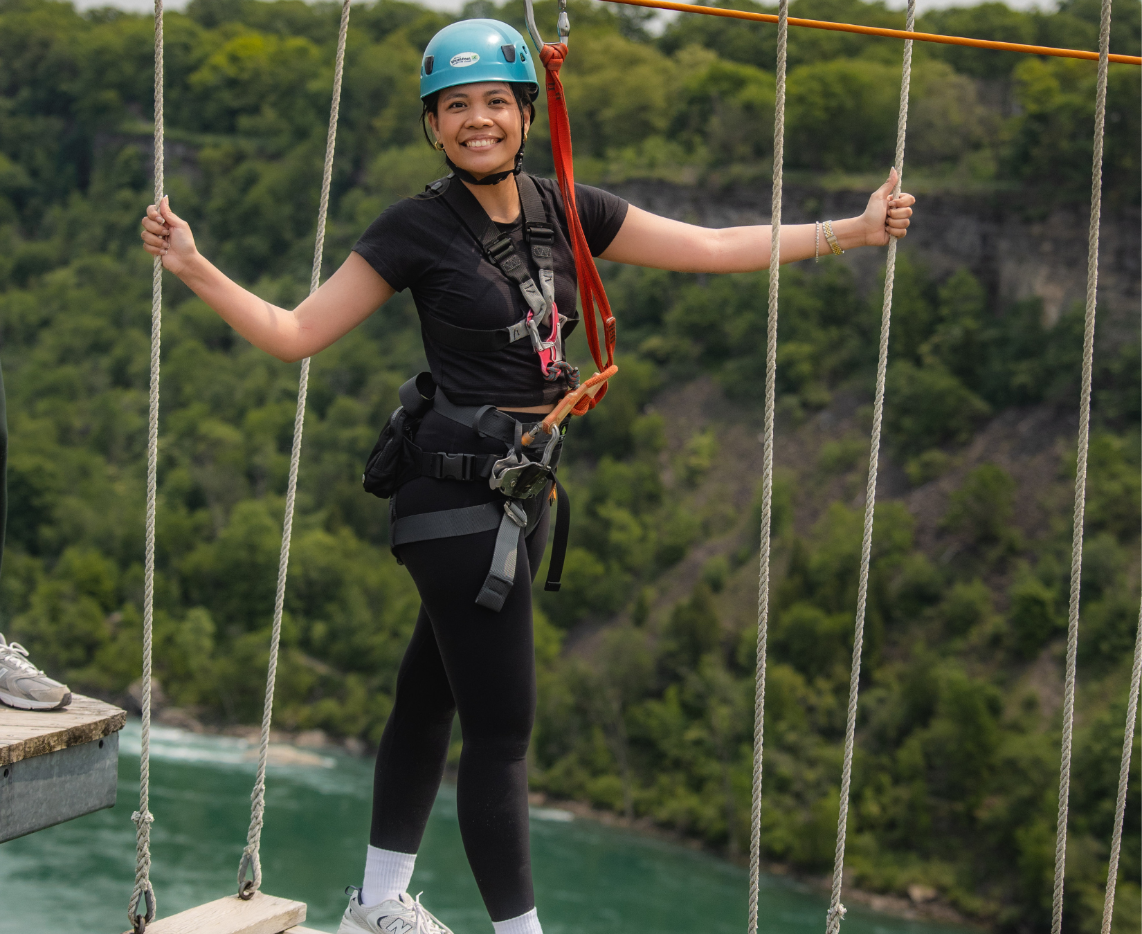 Smiling woman wearing a blue helmet and harness holding ropes while standing on a wooden plank above water with forested hills in the background.