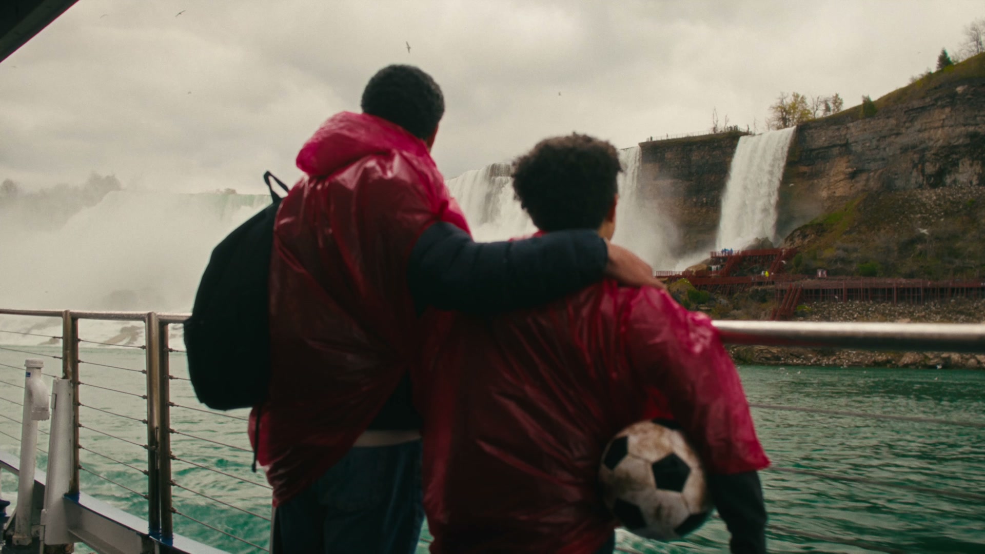 Father and son in rain ponchos looking out over Niagara Falls, soccer ball under arm