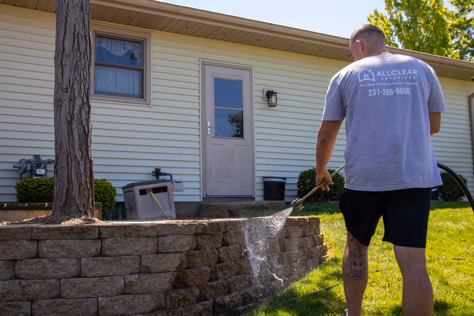 Person using a pressure washer to clean a stone retaining wall outside a house with light siding.