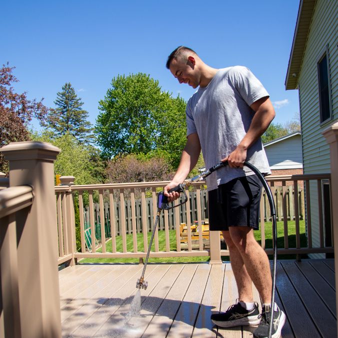 Man using a pressure washer to clean a wooden deck under a clear blue sky.