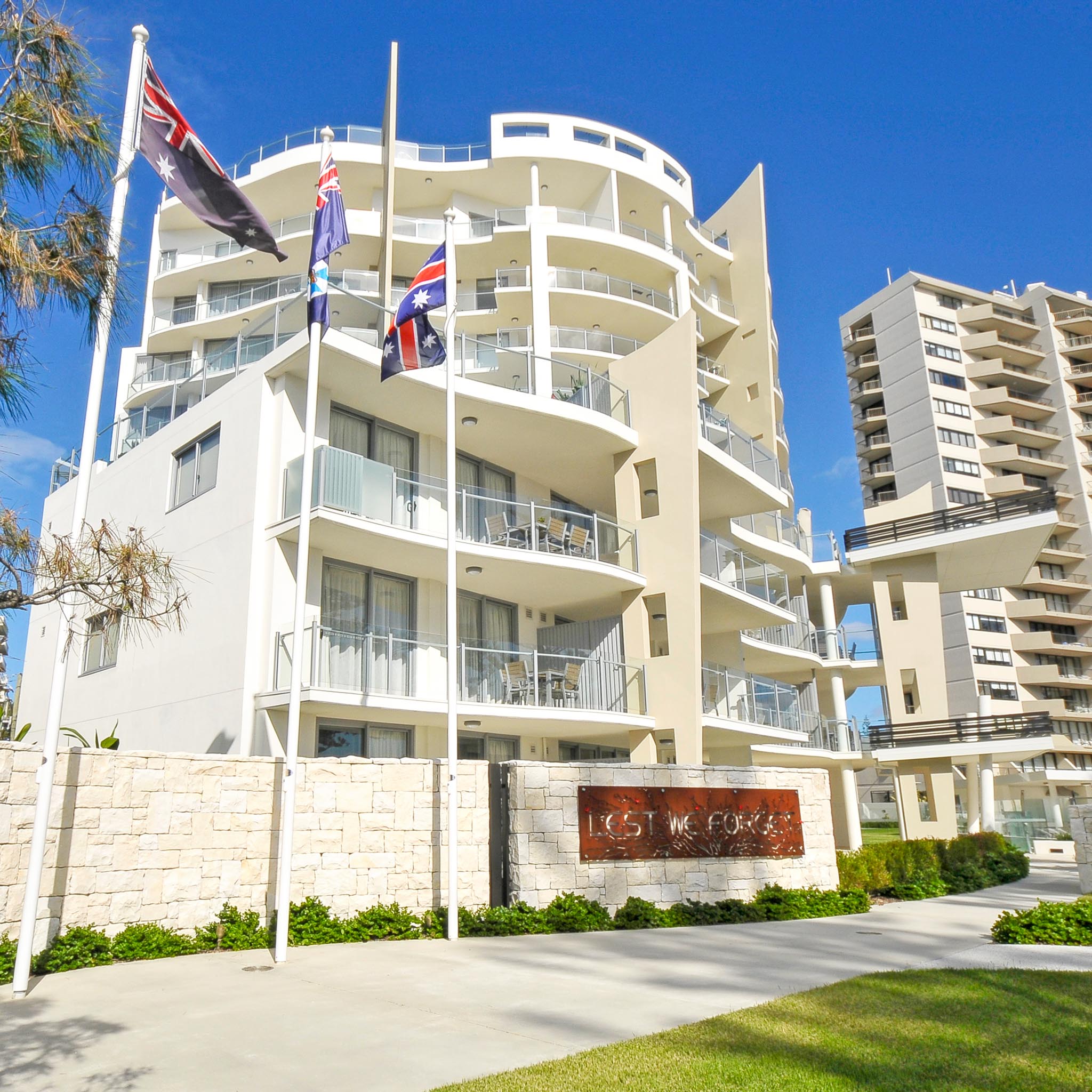 Modern multi-story building with balconies, Australian flags on flagpoles, and a sign reading 'Lest We Forget' on a stone wall.