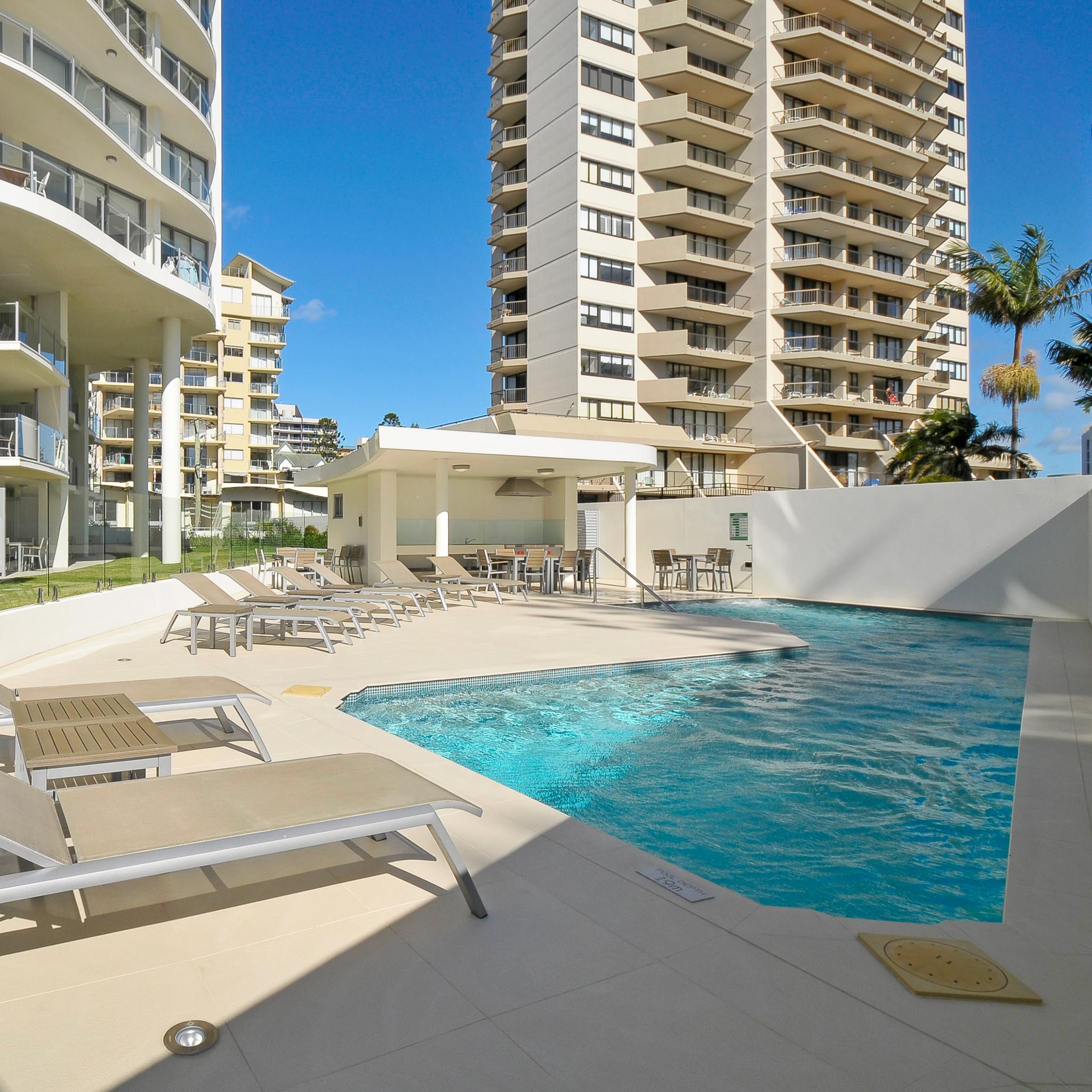 Outdoor swimming pool area with lounge chairs and tables, surrounded by high-rise apartment buildings under a clear blue sky.