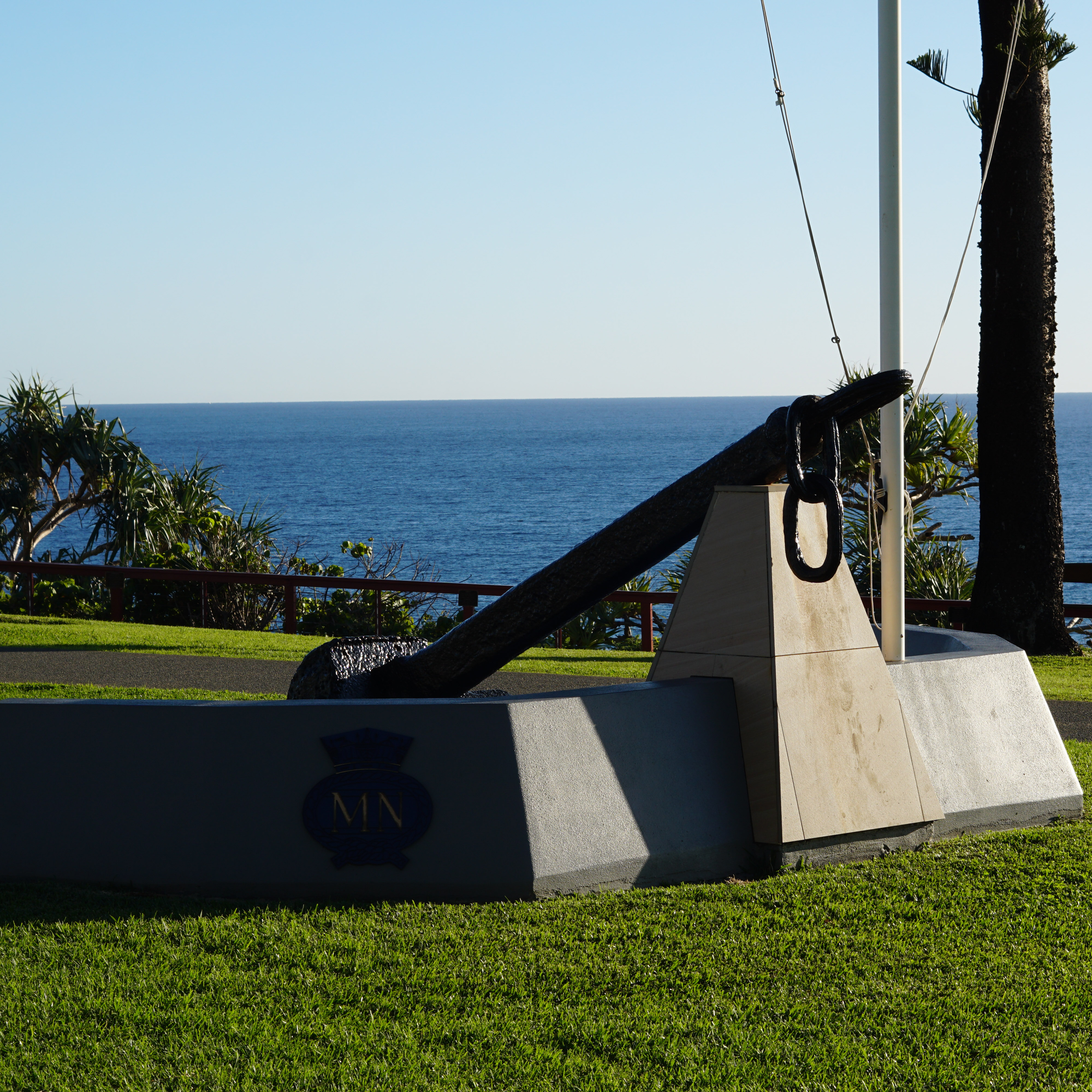 Large black anchor memorial mounted on a stone base with a view of the ocean and grassy lawn.