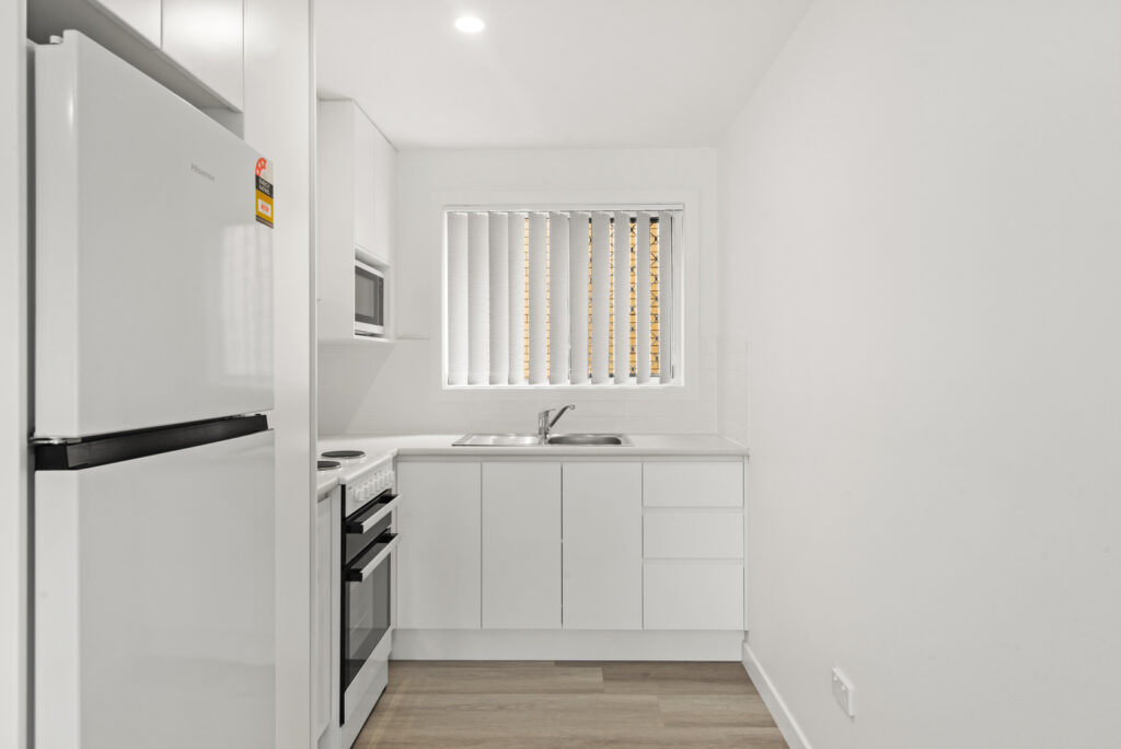 Compact modern kitchen with white cabinets, a refrigerator, stove, microwave, and a window with vertical blinds.