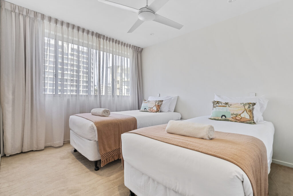 Minimalist bedroom with two single beds, each with white linens, peach throws, and decorative pillows, under a ceiling fan and next to a window with sheer curtains.