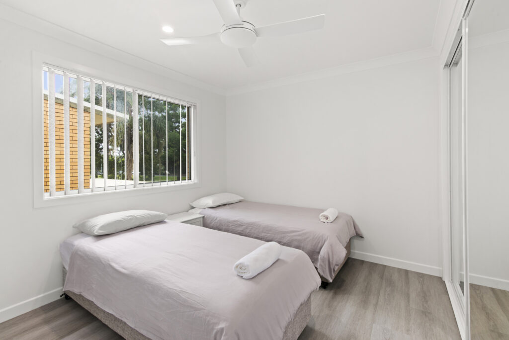 Minimalist bedroom with two single beds, white walls, a ceiling fan, and a large window with vertical bars.