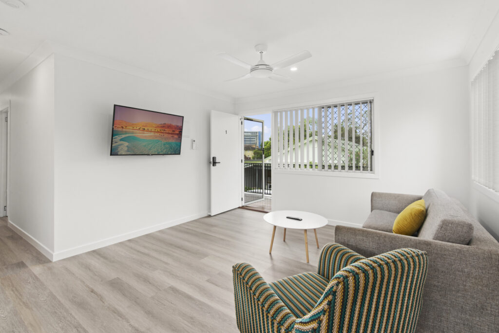Minimalist living room with light wood flooring, a gray sofa with a yellow pillow, a striped armchair, a round white coffee table, wall-mounted TV, ceiling fan, and an open door leading outside.
