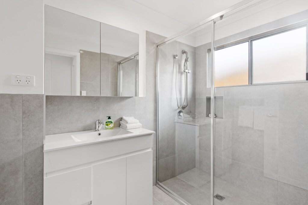 Modern bathroom with white vanity, mirror cabinet, glass-enclosed shower, and gray tiles.