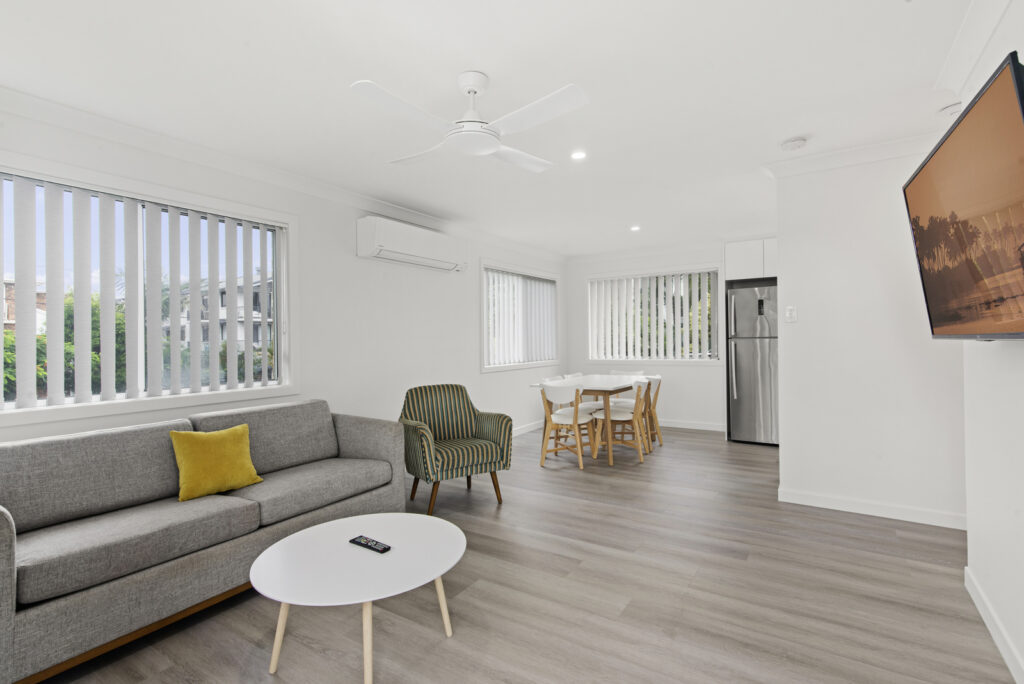Bright living room with a gray sofa including a yellow pillow, striped armchair, white oval coffee table, dining table with chairs, and a wall-mounted TV.