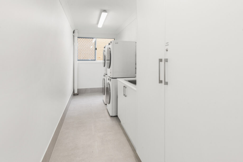 Narrow laundry room with stacked white washer and dryer units, white cabinets, and a window with a security screen.