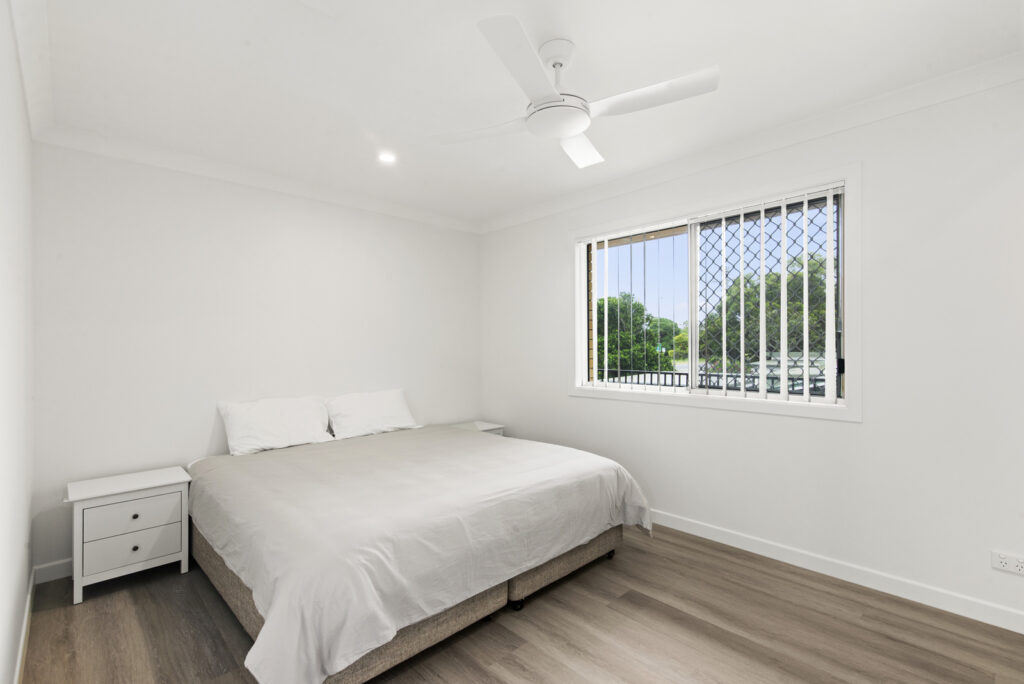 Minimalist bedroom with a large bed, white bedding, bedside table, ceiling fan, and a window with vertical blinds showing greenery outside.