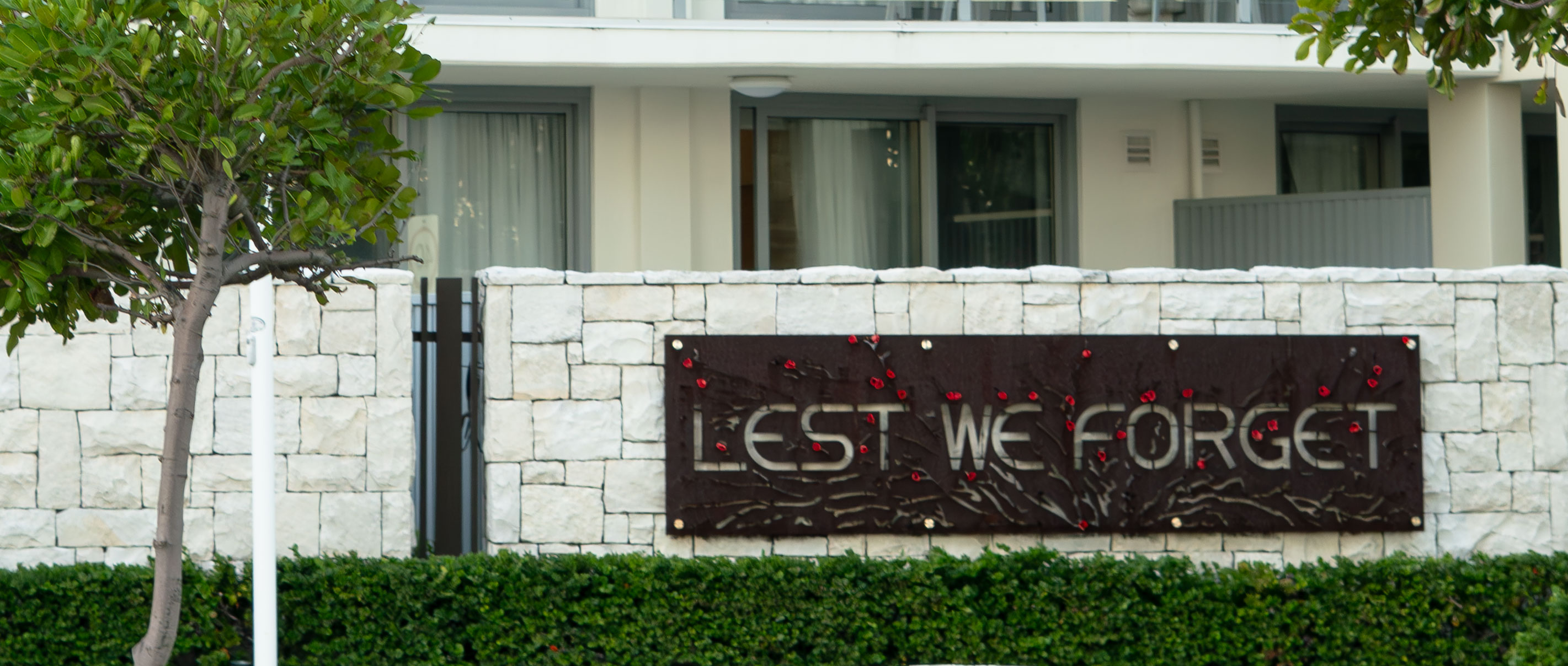 Stone wall with a metal plaque reading 'LEST WE FORGET' decorated with red poppy pins, and greenery in front.