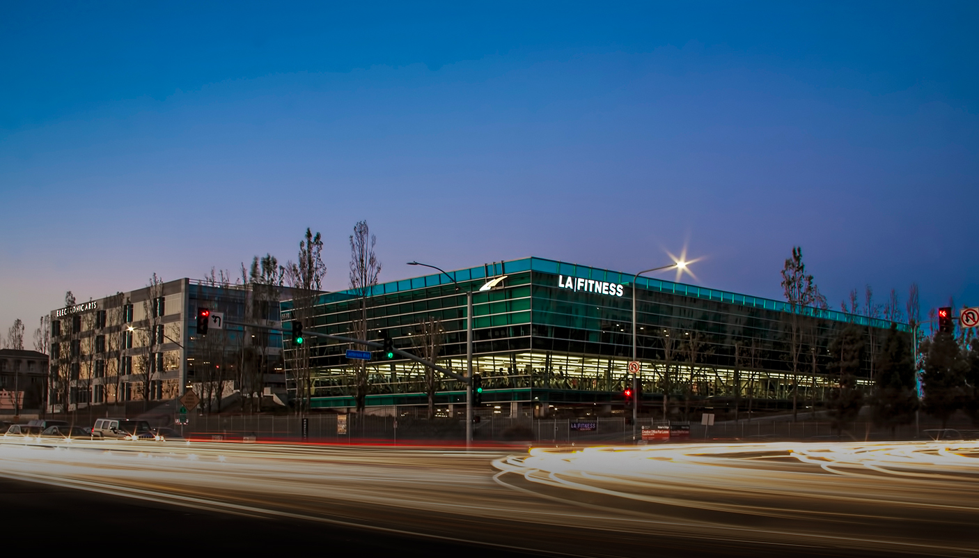 Modern glass LA Fitness building at dusk in Costa Mesa, California, with light trails from passing traffic in the foreground