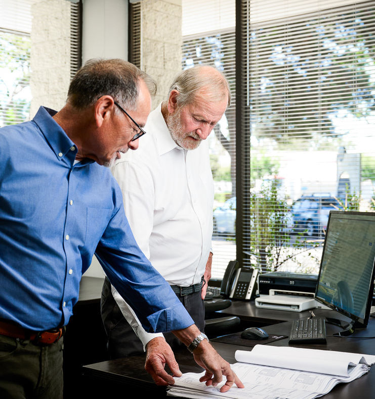 Two senior engineers reviewing architectural and electrical plans together at an office desk with computer and blueprints.