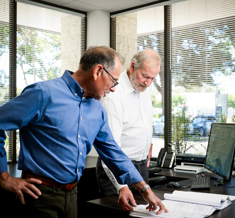 Two senior electrical engineers reviewing blueprints in a sunlit office, one pointing to plans beside a computer workstation