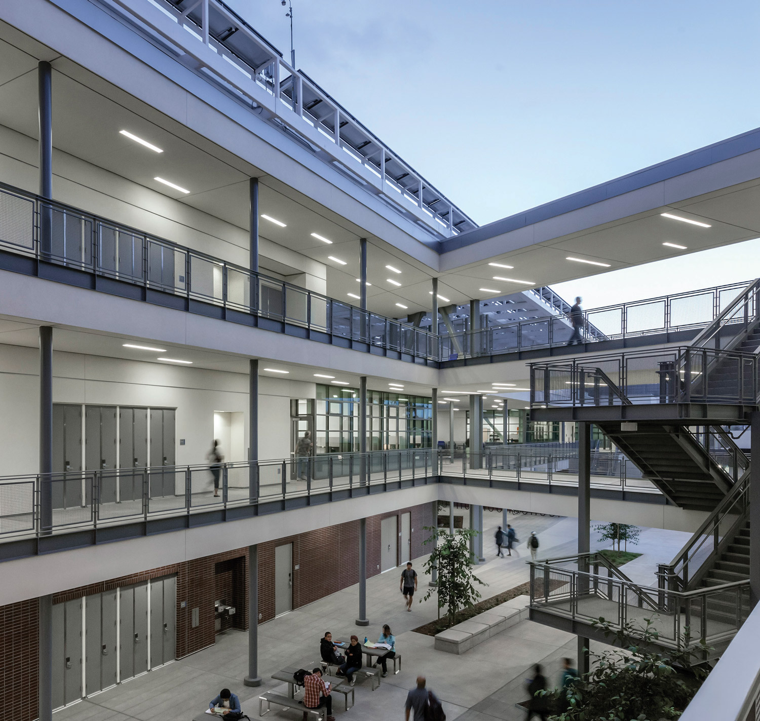 Modern multi-level academic building with open-air courtyard, glass railings, staircases, and students walking and studying outdoors.