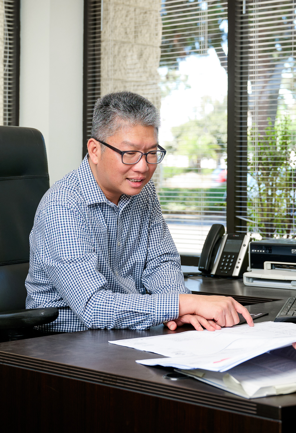 Man in a checkered shirt sitting at an office desk, reviewing documents and pointing while working near a window with blinds.
