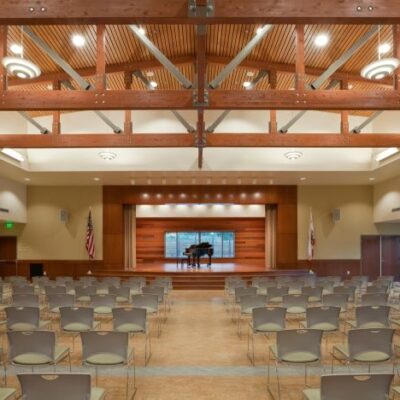 Auditorium interior with stage, wooden ceiling beams, rows of chairs, and piano under warm lighting.