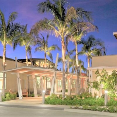 Palm tree-lined walkway with modern covered structures and glowing exterior lighting at night.