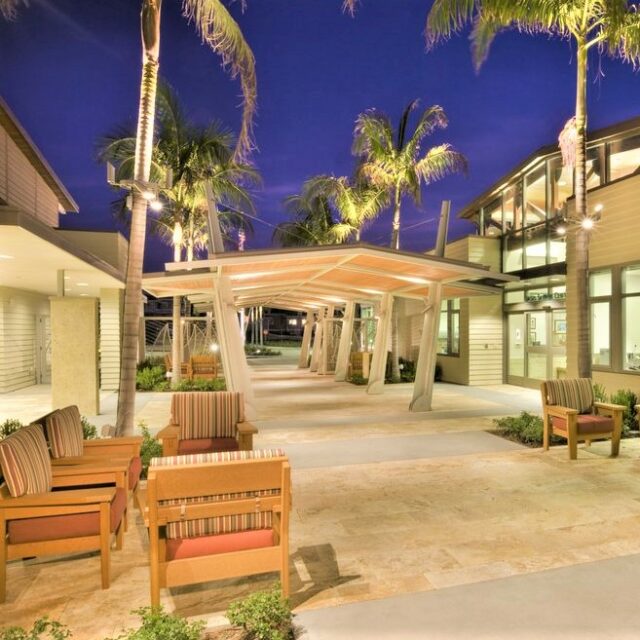 Outdoor seating area with covered walkway, palm trees, and illuminated architecture in the evening.