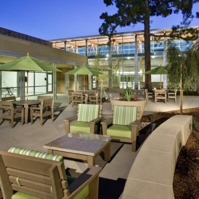 Outdoor seating area at the Fullerton Community Center featuring green cushioned chairs, tables, and umbrellas under evening lighting.