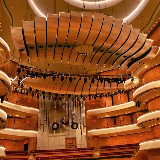 Inside the Segerstrom Concert Hall with sweeping balcony tiers, warm wood finishes, and suspended acoustic panels above the stage.