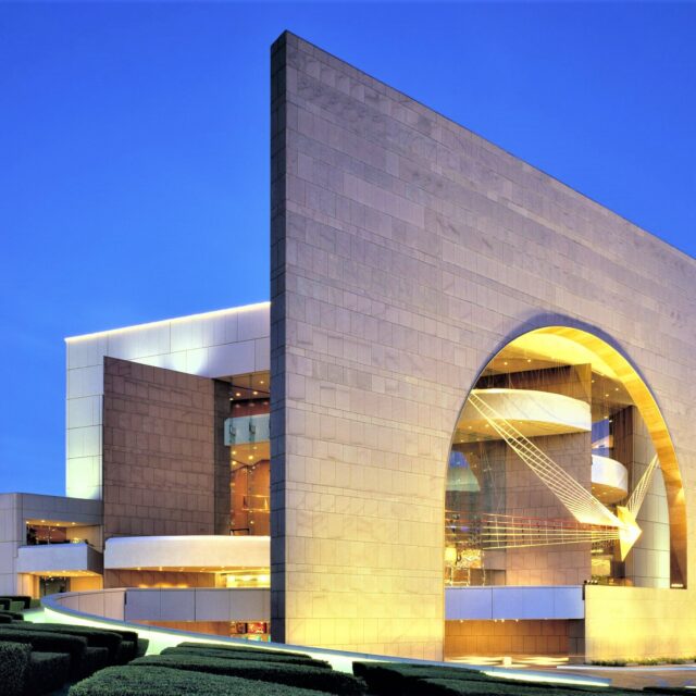 Angled shot of Segerstrom Hall with its monumental stone arch and geometric design glowing under evening lights.