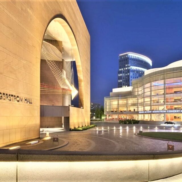 View of the Segerstrom Concert Hall next to Segerstrom Hall, highlighting the dramatic arched entry and surrounding lit plaza at dusk.