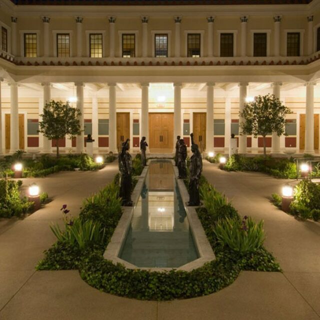 Outdoor courtyard with a central reflecting pool, flanked by bronze statues, greenery, and a colonnade of white pillars.