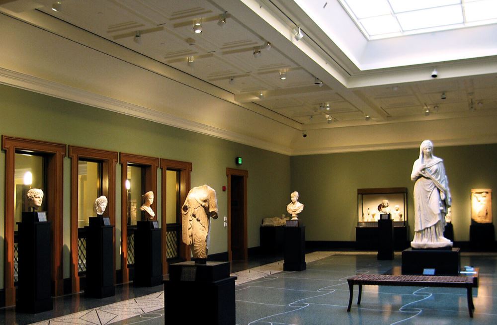 Gallery room with marble statues and busts displayed on pedestals, featuring classical Greek and Roman sculptures under a skylight ceiling.