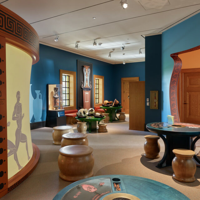 Colorful museum exhibit room with blue walls, interactive tables, stools, and displays focused on ancient Greek pottery and art.