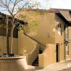 Exterior courtyard view of a two-story beige student housing building with stairways and a tree in the foreground.
