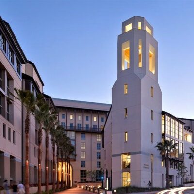 Tall illuminated tower at dusk framed by multi-story housing buildings, lined with palm trees and pedestrian walkways.