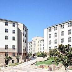 Twin residential buildings with beige facades and black window frames, surrounding a landscaped courtyard with trees and walking paths.