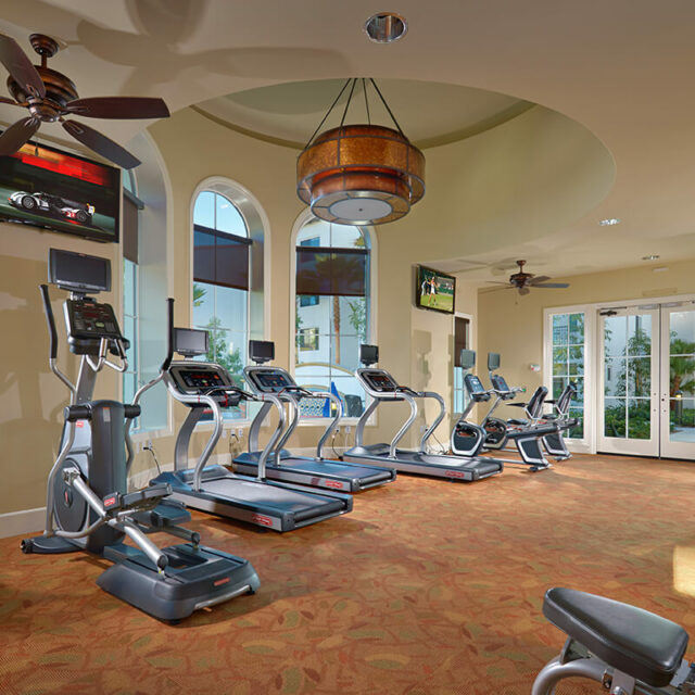 Fitness center with treadmills and exercise bikes facing arched windows, flat-screen TVs, and ceiling fans.