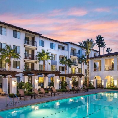Evening view of luxury apartment buildings with balconies overlooking a lit swimming pool and landscaped courtyard.