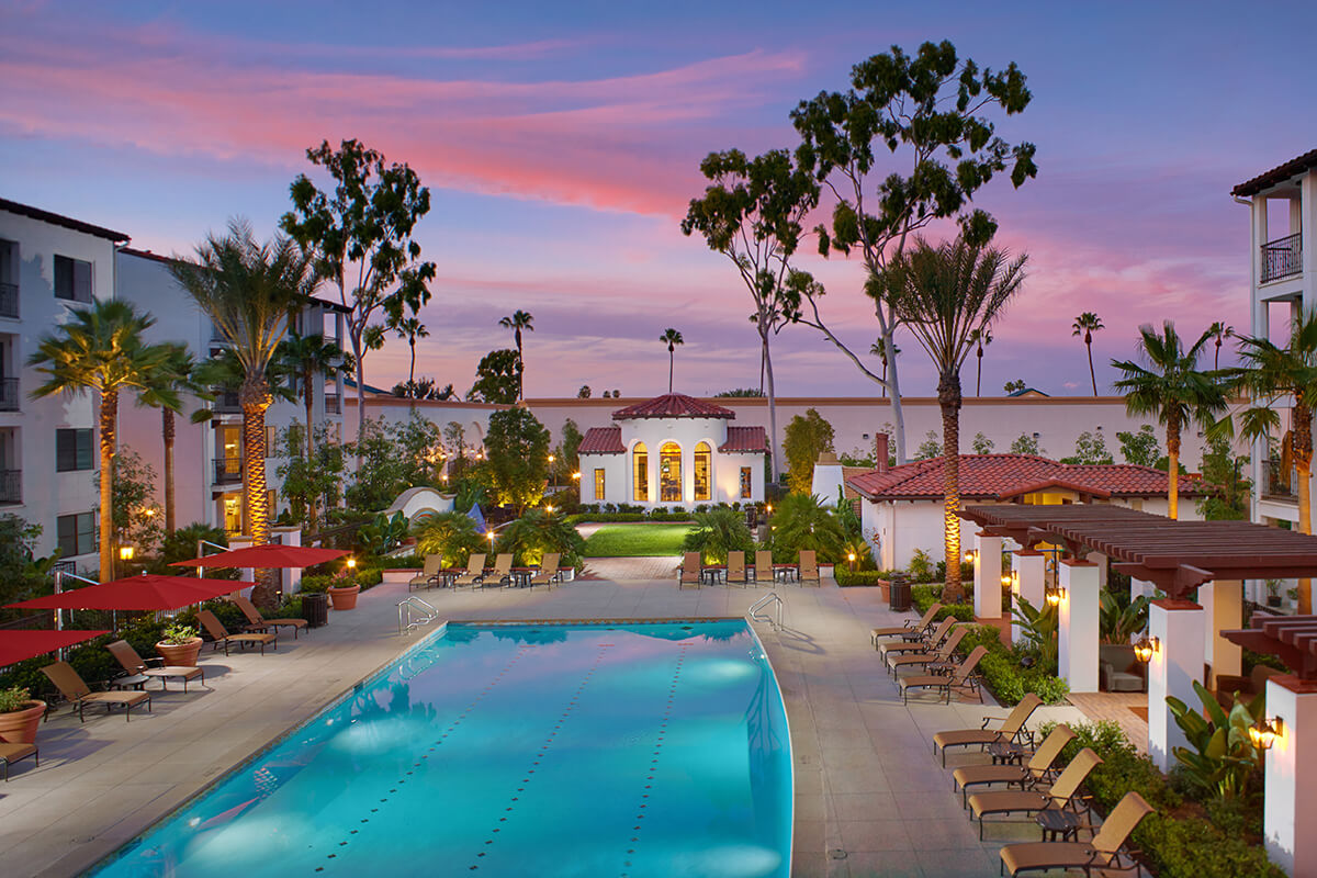 Main entrance of Azulon at Mesa Verde with Spanish-style architecture, red tile roofs, palm trees, and a sign in the foreground.