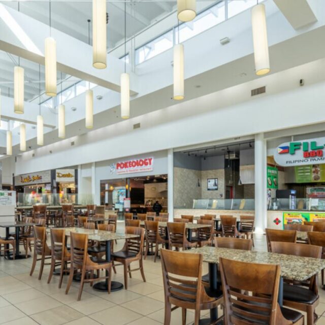 Food court with hanging pendant lights, rows of wooden tables and chairs, and visible restaurant signage.
