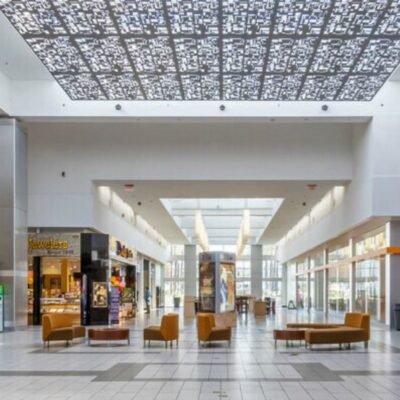 Interior mall corridor with patterned ceiling panels, skylights, storefronts, and modern seating.