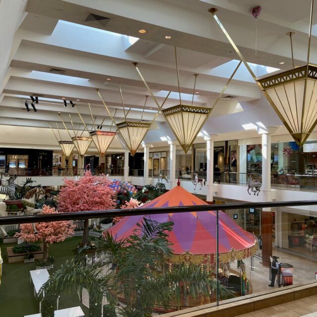Interior atrium of South Coast Plaza with carousel, decorative lighting fixtures, pink cherry blossom trees, and upper-level retail walkways.