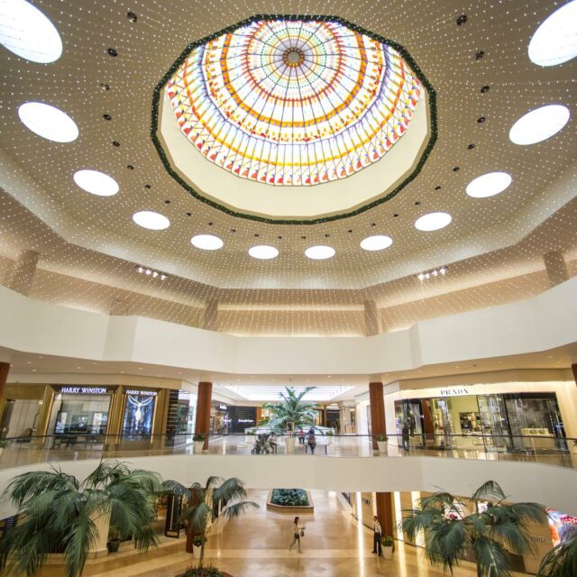 Luxury retail hall featuring a grand stained-glass dome ceiling, decorative lighting, palm trees, and high-end shops.