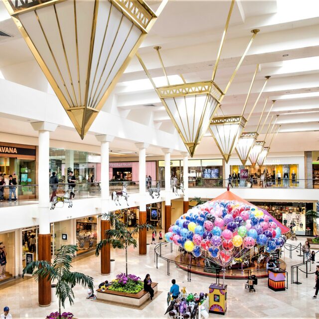 Interior view of South Coast Plaza with two levels of retail shops, oversized hanging light fixtures, and a colorful balloon display in the central atrium.