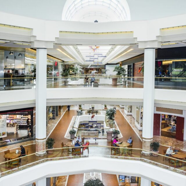 Multi-level indoor shopping mall with open atrium, skylight ceiling, retail storefronts, and seating areas.