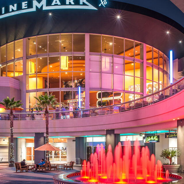 Cinemark movie theater entrance with large curved glass facade, interior lights glowing, and a red-lit fountain in front.