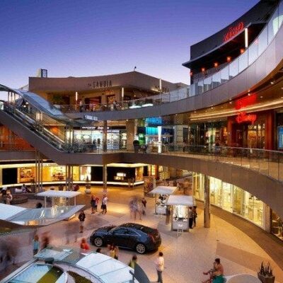 Multi-level open-air shopping and dining area at night, with escalators, modern storefronts, and crowds of visitors.