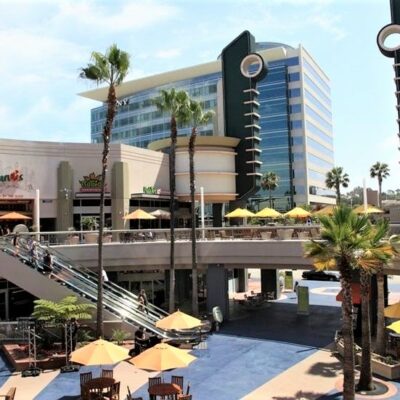 Outdoor retail and dining plaza with escalators, palm trees, and yellow umbrellas, adjacent to a modern office tower.